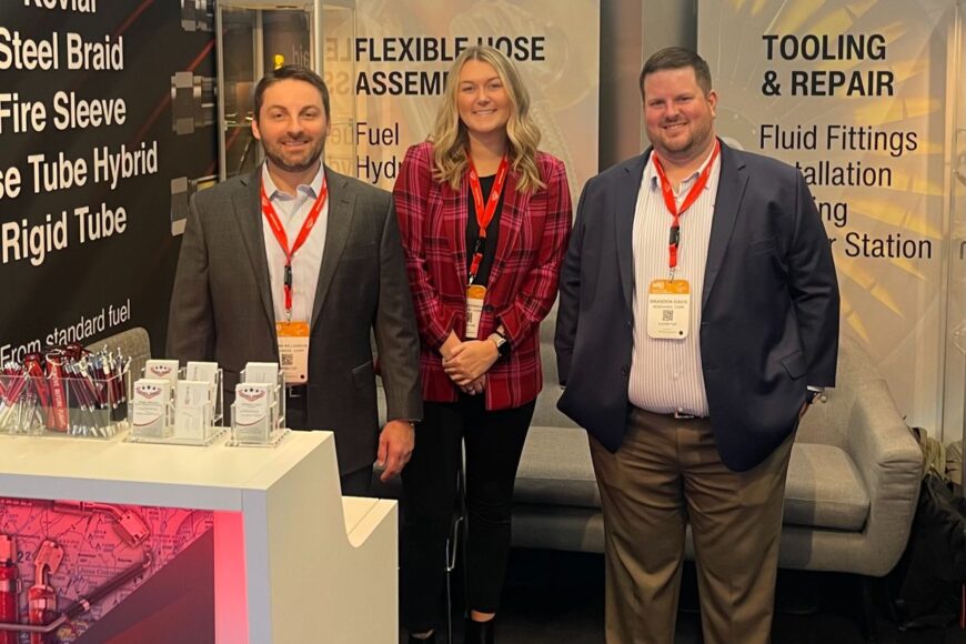three professionals standing at a trade show booth featuring hoses and aerospace equipment, smiling for the photo with seo materials.