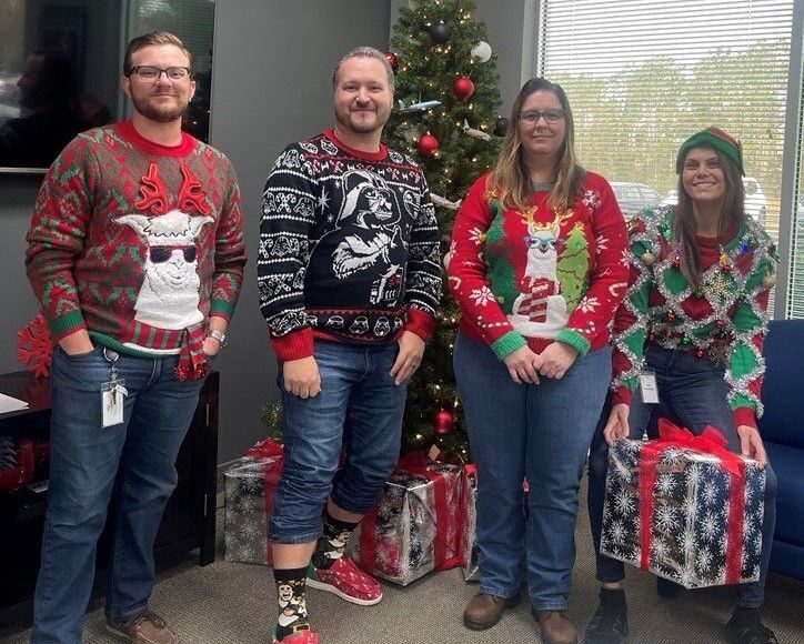 four coworkers in festive sweaters posing in an office with a christmas tree and gifts in the background.