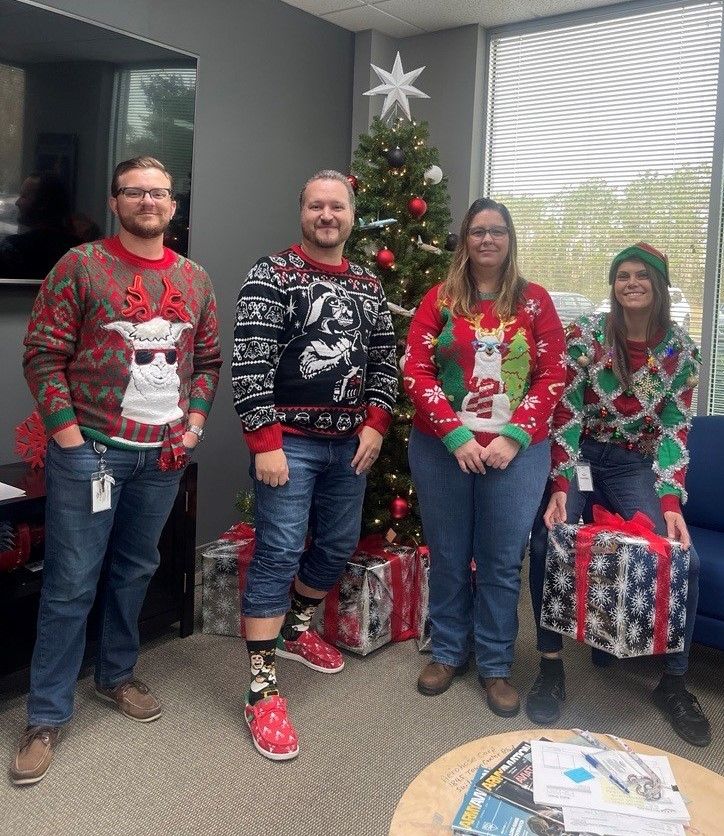 four coworkers in festive sweaters posing in an office with a christmas tree and gifts in the background.