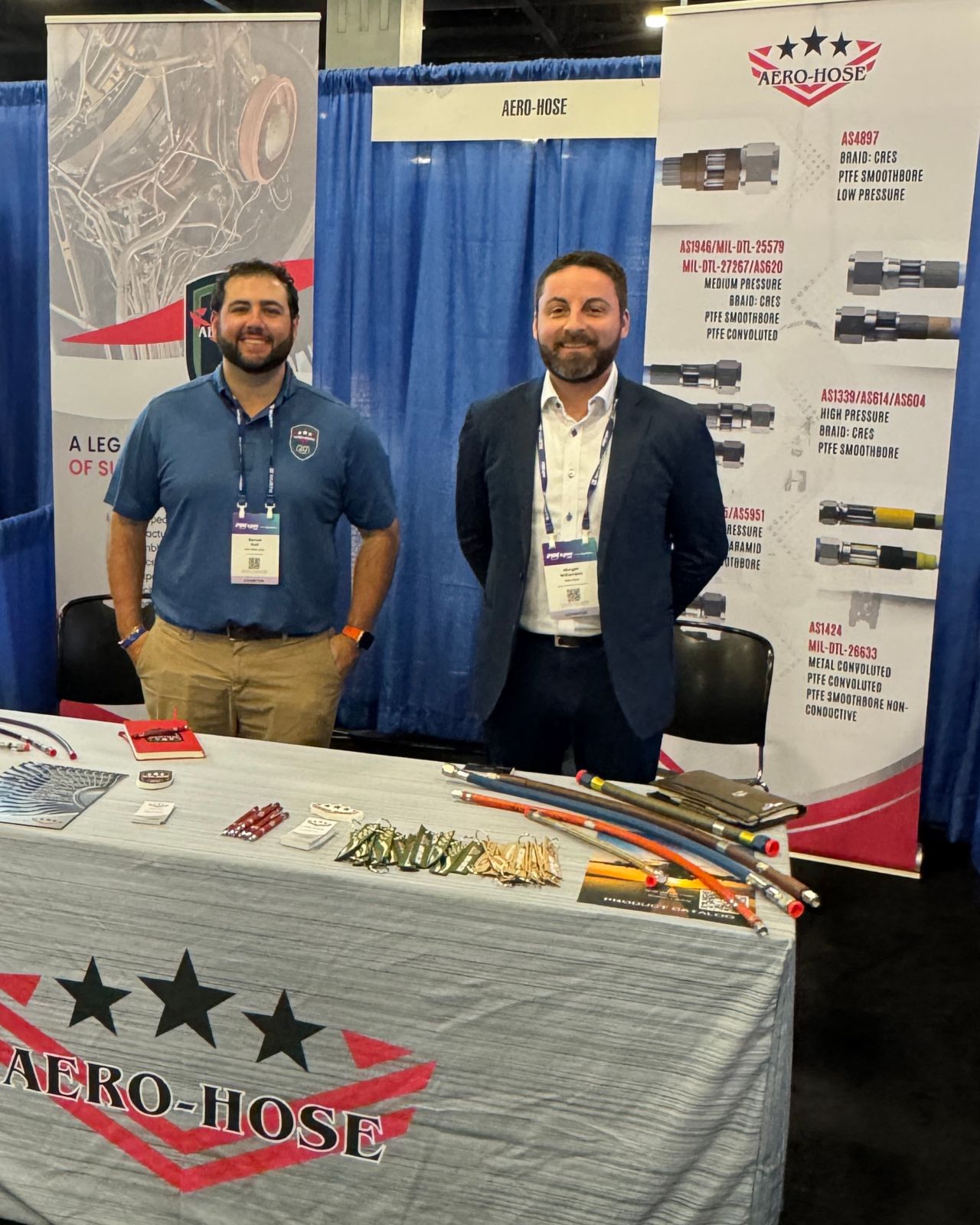 Two people standing behind a trade show booth displaying Aero-Hose products and promotional materials.