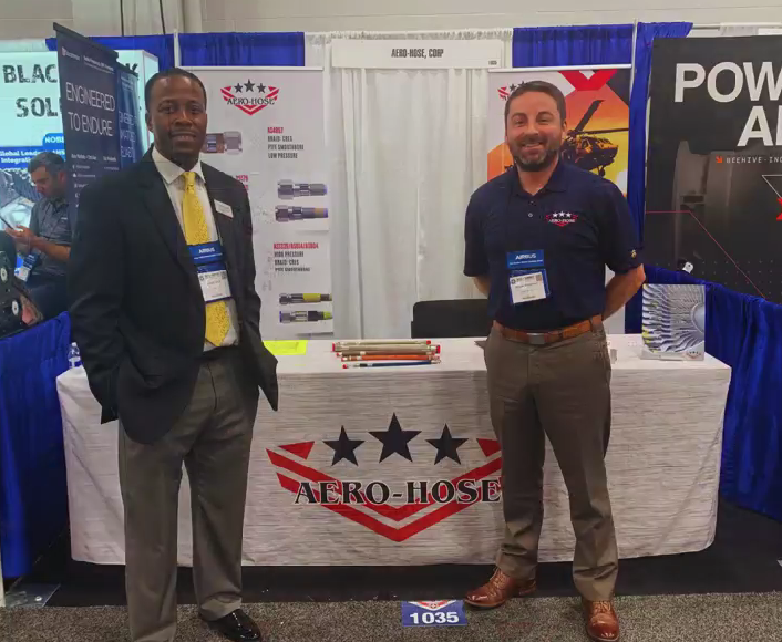 Two men stand in front of an Aero-Hose booth at a trade show, with promotional materials and banners displayed behind a table draped with the company's logo.